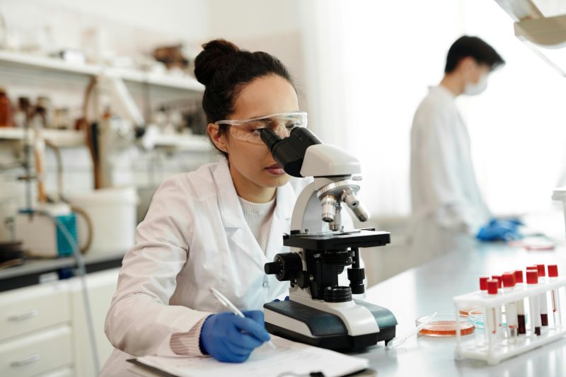Woman scientist examining samples under a microscope in a laboratory setting, symbolizing medical research and disease investigation.